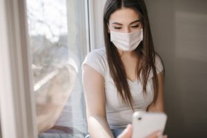Attractive woman in medical mask sits on windowsill at home. Female in white t'shirt and white medical mask use phone